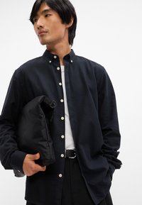 Cămașă button-up de un albastru închis cu guler mandarin, având butoane deschise și mâneci lungi, asortată cu o geantă neagră căptușită.