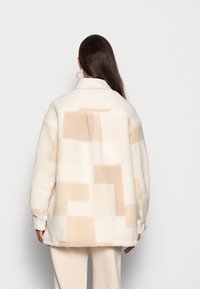 Fleece jack in ivoorkleur met beige geometrische patronen, knoopmanchetten en een kraag. Zachte textuur met een losse pasvorm.