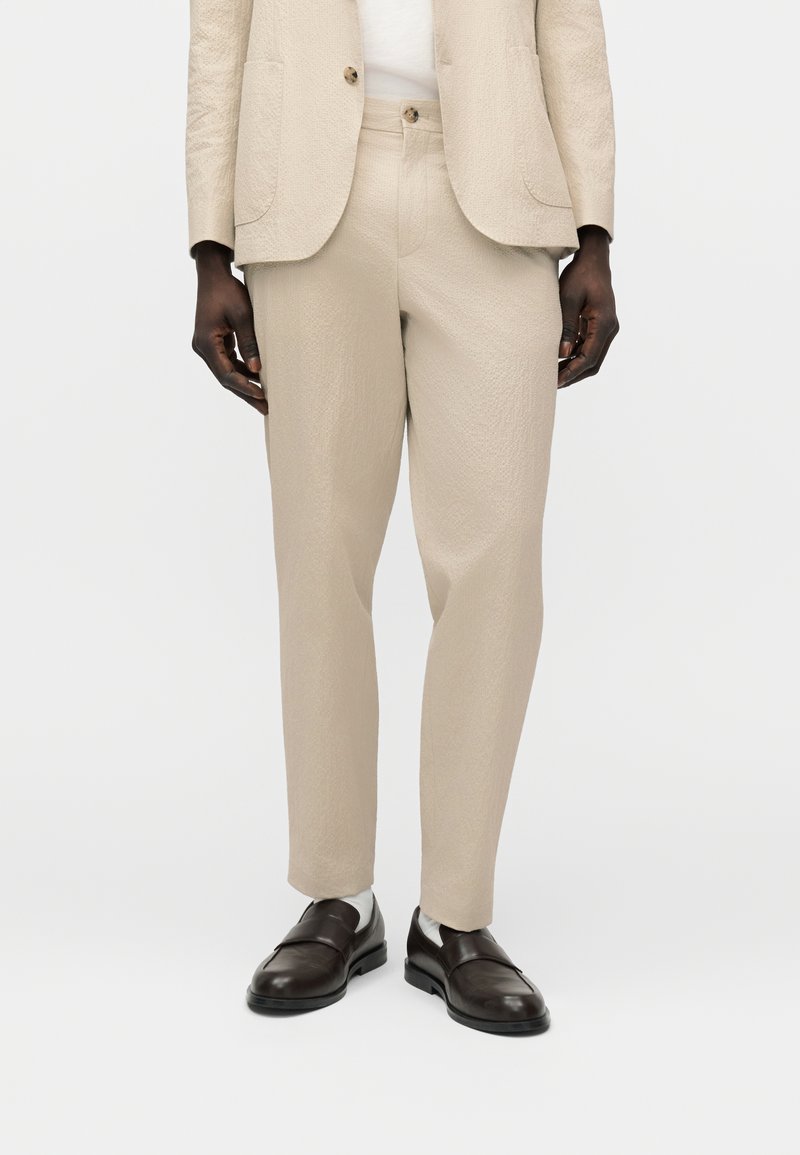 Άνδρας με μπεζ υφασμάτινο παντελόνι και σακάκι, λευκό πουκάμισο, λευκές κάλτσες και μαύρα δερμάτινα μοκασίνια, στέκεται απέναντι σε λευκό φόντο.