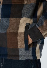 Fliisist kangast särk-jakk ruudulise mustriga pruunis, sinises ja kreemjas toonis. Omab nööbivarrast ja küljetaskut kontrastse riidest äärega.