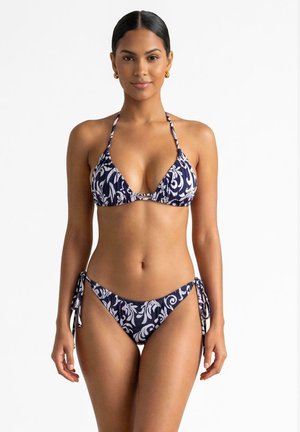 Kobieta ubrana w granatowy i biały wzorzysty bikini na szyję z wiązanymi z boku majtkami, stojąca na jednolitym białym tle.