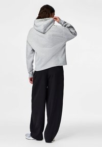 Henkilö, jolla on pitkät hiukset, pukeutunut vaaleanharmaaseen huppariin ja löysiin mustiin housuihin, säätää hupun paikkaa, seisoo valkoisella taustalla.