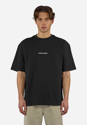 Nuori aaltoilevatukkainen mies pukeutuneena mustaan Pegador T-paitaan ja beigeihin housuihin, seisoo yksinkertaisen valkoisen taustan edessä, näkyvissä pieni ristitatuointi.