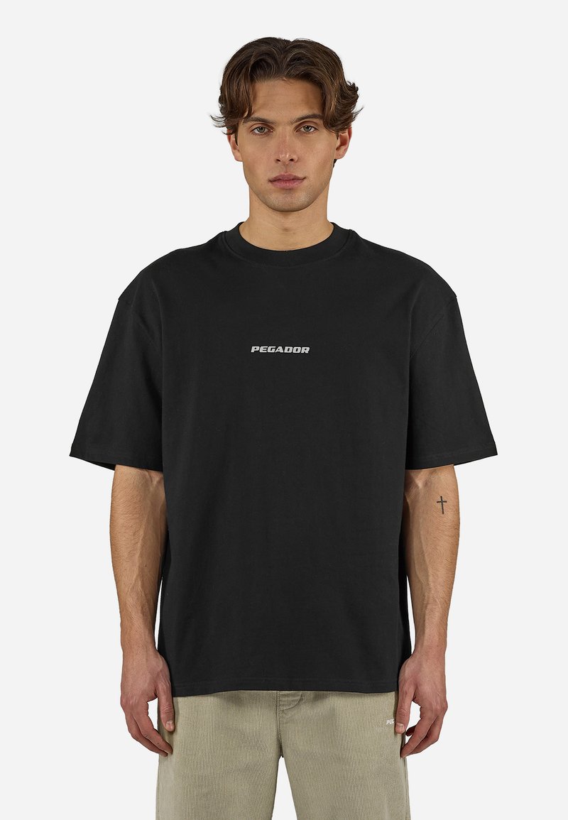 Νέος άνδρας με κυματιστά μαλλιά φοράει μαύρο T-shirt Pegador και μπεζ παντελόνι, στέκεται μπροστά σε απλό λευκό φόντο με ένα μικρό τατουάζ σταυρού ορατό.