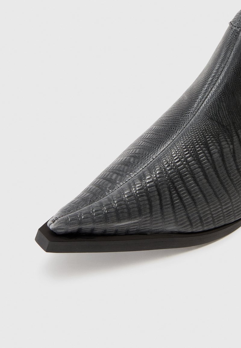 Bottine à bout pointu en matériau texturé gris foncé. Présente une silhouette élégante avec une semelle noire plate et des détails en relief discrets.