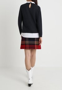 Kvinna iförd svart långärmad tröja över vit skjorta, röd rutmönstrad kjol och vita stövlar som går till mitten av vaden, stående vänd bort från kameran mot en enfärgad bakgrund.