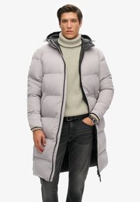 Abrigo largo acolchado en gris claro con cremallera negra y capucha, usado sobre un cuello de tortuga texturizado en crema, combinado con jeans oscuros.