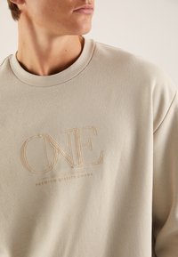 Beige sweatshirt with a crew neck, featuring raised embroidered text "ONE" and "PREMIUM QUALITY GOODS" in light tones on the front.