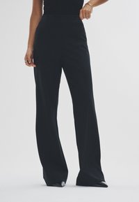 Sorte bukser med brede ben i et glat, struktureret stof. Høj talje, sidesømme og et rent snit, kombineret med spidse sorte sko.