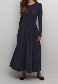 Navyblauwe jurk met lange mouwen, een geribbelde bovenkant en een geplooide rok. Zachte stof met een vloeiende silhouet en platte, zwarte schoenen zichtbaar.