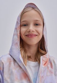 Leichte, pastellfarbene Batikjacke mit Kapuze und Reißverschluss. Verfügt über weiße Reißverschlussdetails und weichen Stoff. Darunter ist ein schlichtes weißes Shirt sichtbar.