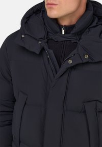 Tmavě námořnická péřová bunda s vysokým límcem, odnímatelnou kapucí a zipovým zapínáním. Má zaklapávací uzávěry a dvě boční kapsy.