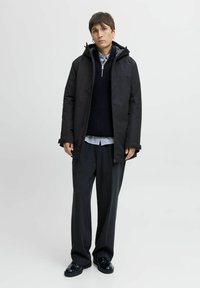 Sort yderjakke med hætte, lagd ovenpå en mørk sweater og en stribet skjorte. Løse grå bukser og sorte sko fuldender outfittet.