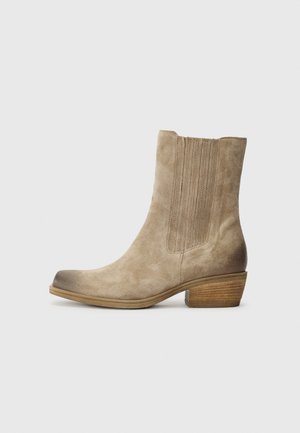 Beige suede enkellaarsjes met elastische zijpanelen, puntige neus en een gestapelde houten hak. Gladde textuur met subtiele schaduw aan de randen.