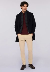 Tmavý kabát přes černou prošívanou vestu, vínové polo, světle béžové kalhoty a hnědé boty. Neformální vrstvení s strukturovaným designem.