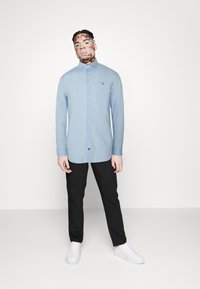 Hellblaues Langarmshirt mit Button-Down-Kragen und dezentem Logo auf der Brust, kombiniert mit schwarzen Hosen und weißen Sneakers.