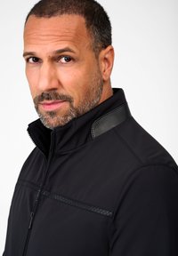 Schwarze Jacke mit Stehkragen und Lederakzenten an den Schultern. Glatte Textur mit Reißverschluss und dekorativem Stitching.