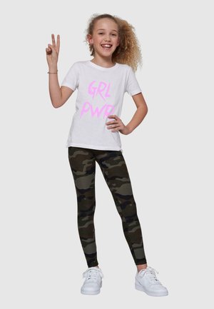 T-shirt blanche avec le texte rose "GRL PWR", associée à des leggings camouflés verts. La mannequin porte des baskets blanches et pose avec un signe de paix.