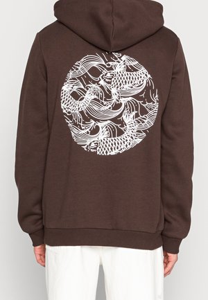 Brauner Hoodie, getragen von einer Person mit dem Rücken zur Kamera, mit weißem kreisförmigem Koi-Fisch- und Wellendesign auf dem Rücken, kombiniert mit weißen Hosen.