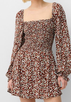 Femme portant une mini-robe smockée à manches longues, fleurie, avec un col carré dans des tons marron, blanc et rouge.