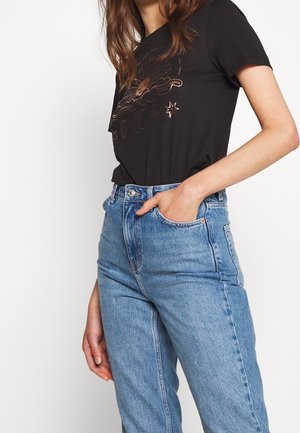 Kobieta ubrana w czarny t-shirt z wzorem słońca i gwiazd, wpuszczony w niebieskie jeansy z wysokim stanem, z jedną ręką w przedniej kieszeni.