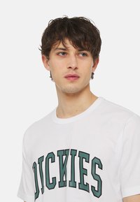 Bílé bavlněné tričko s výraznou zelenou grafikou "DICKIES" v klenutém písmu přes hruď. Klasický kulatý výstřih a krátké rukávy.