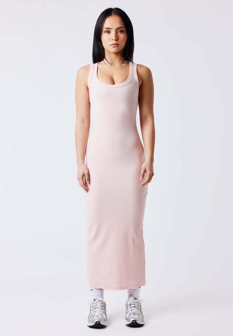Femme debout de face portant une robe maxi sans manches côtelée rose clair, des chaussettes blanches et des chaussures de sport blanches sur un fond uni.
