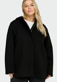 Manteau noir à capuche avec une coupe décontractée, une texture lisse et une fermeture éclair frontale. Comporte des manches larges et un design minimaliste.