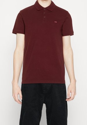 Hombre que lleva una camisa polo de manga corta color rojo oscuro con tres botones y un pequeño logo en el pecho, combinada con pantalones de pana negros.