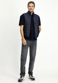 Granatowa kamizelka puchowa z wysokim kołnierzem, noszona na czarnej koszuli i szarych dżinsach. Szare sneakersy z czarnymi akcentami dopełniają stylizację.