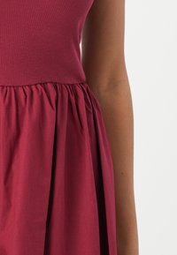 Robe bourgogne avec corsage côtelé, taille froncée et jupe lisse et fluide. Le tissu a une texture douce, idéale pour un usage quotidien.