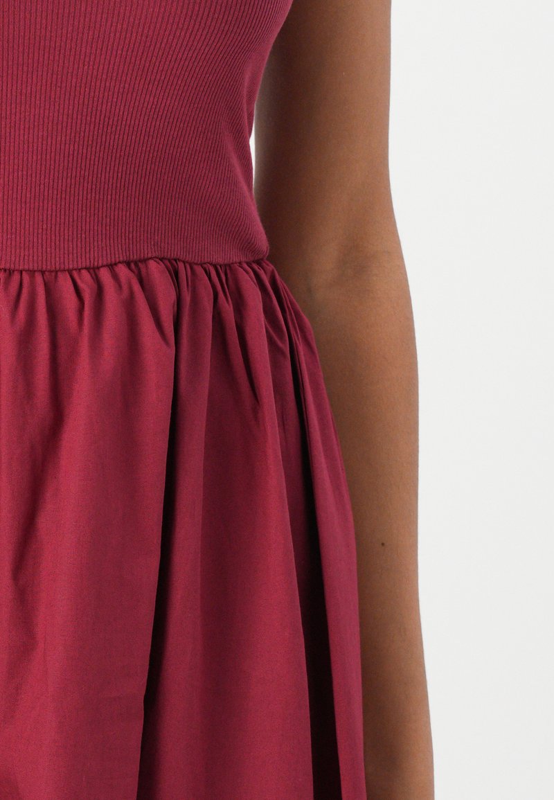Robe bourgogne avec corsage côtelé, taille froncée et jupe lisse et fluide. Le tissu a une texture douce, idéale pour un usage quotidien.
