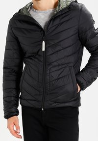 Svart puffjacka med zippered fram, horisontell sömnad och grönt mönstrat foder inuti huvan. Har sidofickor.