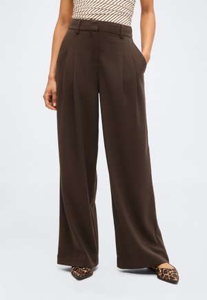 Γυναίκα που φοράει παντελόνι καφέ σκούρο με φαρδιά μπατζάκια και πιέτες, και μυτερά παπούτσια με λεοπάρ σχέδιο, στέκεται με το ένα χέρι στην τσέπη.