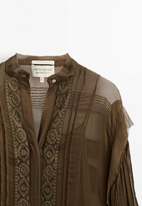 Genomskinlig brun blus med vertikala veck, prydd halsringning och dekorativ broderi, med en mandarin-kräg och knapplås.