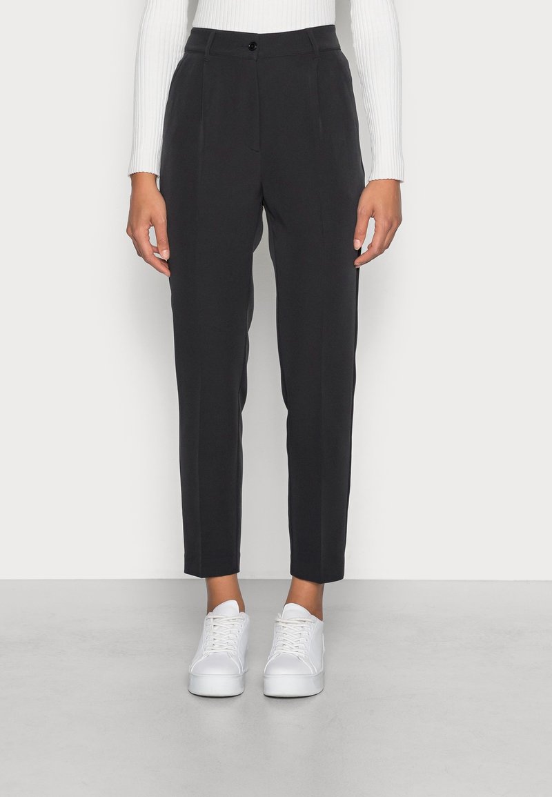 Femme portant un pantalon noir court et ajusté avec des plis sur le devant, un haut blanc côtelé à manches longues, et des baskets blanches, debout sur un sol gris.