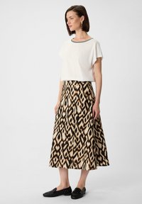 Weißes Kurzarmoberteil mit schwarzem Saum, kombiniert mit einem langen, gemusterten Rock, der braune, beige und schwarze geometrische Designs aufweist. Schwarze Schuhe.