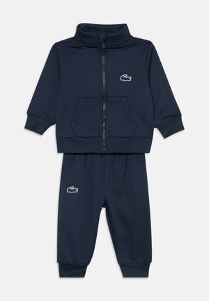 Survêtement bleu marine pour tout-petit avec veste zippée devant et pantalon à taille élastique, tous deux ornés d'un logo crocodile blanc sur la poitrine et la jambe.