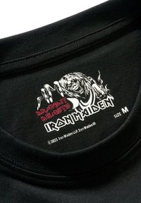 Juodas medvilninis marškinėlių apykaklė su raudona ir balta grafika, kurioje yra tekstas "BRANDIT BEASTS IRON MAIDEN" ir braukiančios rankos iliustracija.