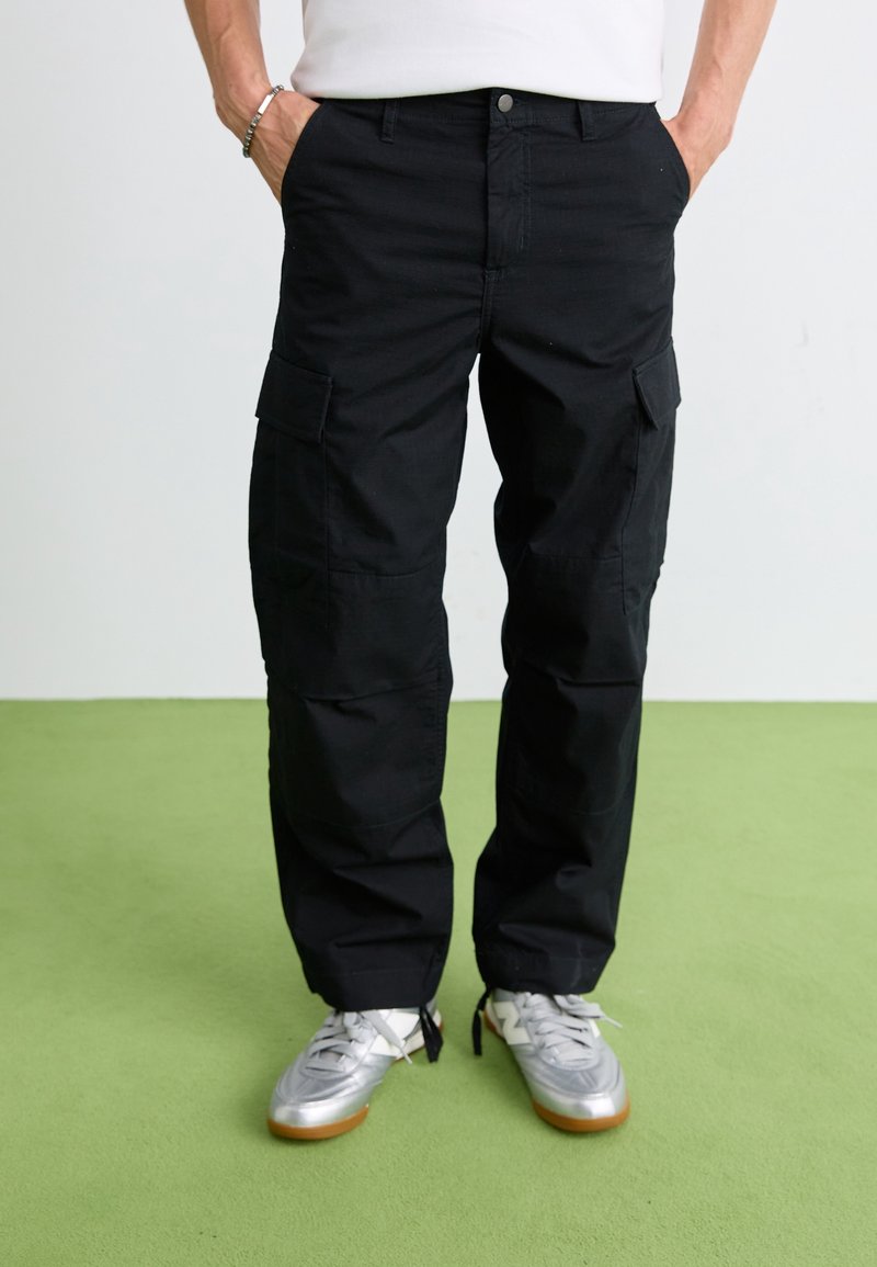 Czarne spodnie cargo wykonane z wytrzymałej tkaniny, z wieloma bocznymi kieszeniami, luźnym krojem i zapinane na guzik. Noszone w połączeniu ze srebrnymi butami sportowymi.