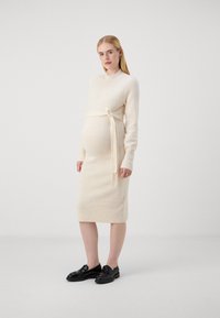 Abito lavorato a maglia color crema con maniche lunghe, scollatura rotonda e una cintura in vita. Indossato con mocassini neri. Tessuto morbido, vestibilità aderente.
