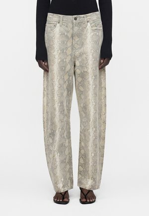 Personne portant un pantalon large beige et gris à motif peau de serpent, des sandales à talons noires et un haut noir à manches longues.