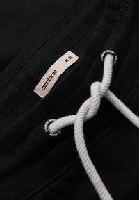 Schwarzer Stoff mit weißem Kordelzug, der durch gebrandete Ösen geführt wird, und einem beigen Etikett mit der Aufschrift "ombre" und Größe "M EUR 48".