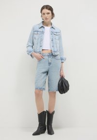 Giacca di jeans azzurro chiaro, top corto bianco, pantaloni denim abbinati, borsa in pelle nera e stivali cowboy neri a punta. Insieme casual con texture distinte.