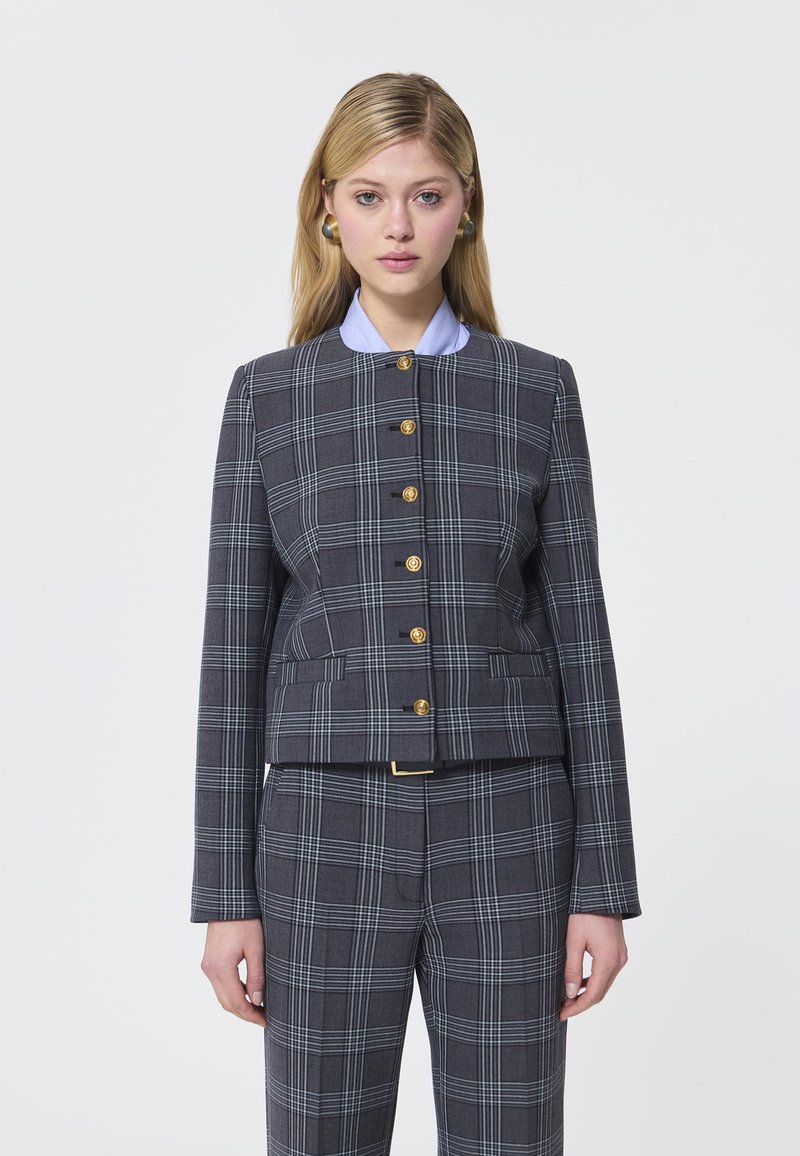 Blazer gri cu carouri, cu nasturi aurii, croială ajustată, având un guler rotund peste o cămașă bleu deschis. Mâneci drepte și buzunare laterale.