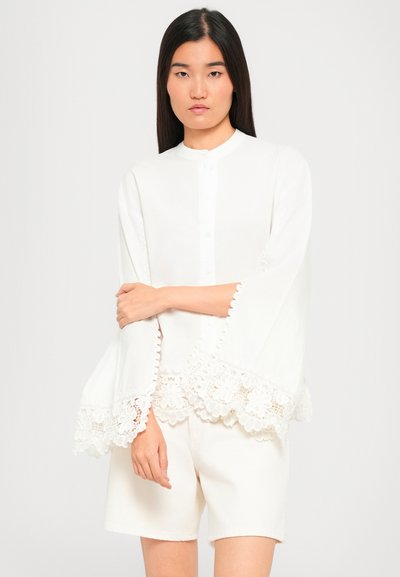 Young woman wearing a white blouse with wide lace-trimmed sleeves and white shorts, standing against a plain light background.