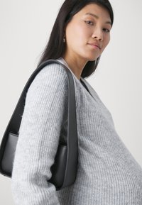 Sac à épaule en cuir noir avec un design structuré, doté d'une texture lisse et d'une large bandoulière. Le modèle porte un pull en tricot gris clair.