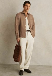 Cămașă button-up de culoare maro deschis cu un buzunar, pantaloni crem și o geantă tote texturată de culoare maro. Pantofii de culoare maro închis completează ținuta.