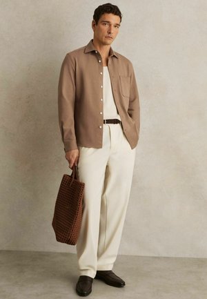 Cămașă button-up de culoare maro deschis cu un buzunar, pantaloni crem și o geantă tote texturată de culoare maro. Pantofii de culoare maro închis completează ținuta.