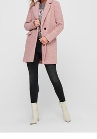 Rosa strukturerad kappa med snedskurna slag, knappstängning med en knapp och sidfickor, kombinerad med svarta tajta jeans och krämfärgade ankelboots.